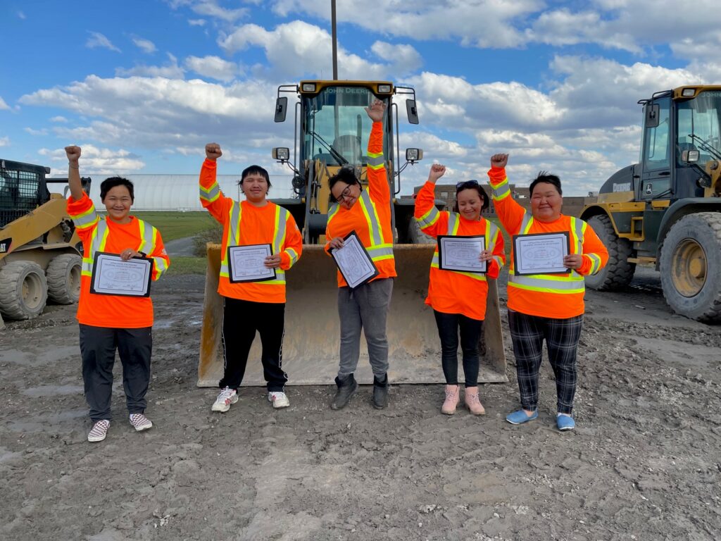 5 successful graduates of the Heavy Equipment Operators program in bright orange safety equipment with their fists in the air and holding their certificates while standing in front of a bull dozer.