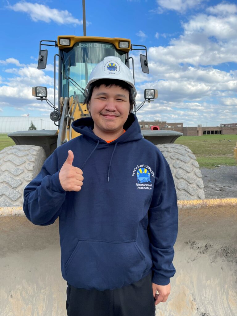Student standing infront of a heavy equipment maching wearing a QIA hardhat and sweater smiling with a thumbs up.