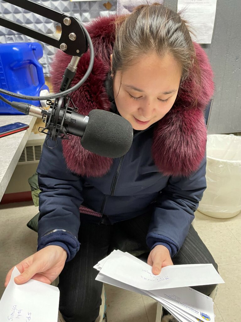 Woman sitting down in front of a microphone looking at envelopes smiling and reading the names out.