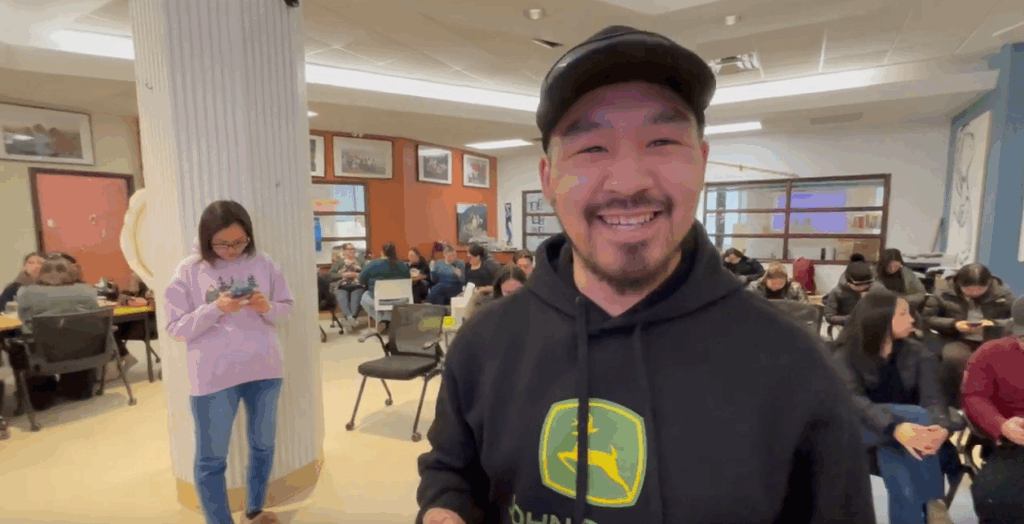 A tuttarvik app user is smiling widely at the camera, while people are sitting at tables behind him. A woman can be seen in the background looking at her phone.