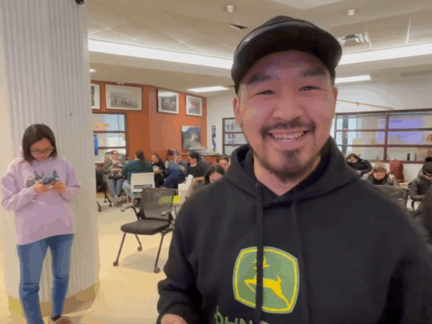 A tuttarvik app user is smiling widely at the camera, while people are sitting at tables behind him. A woman can be seen in the background looking at her phone.